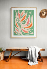Abstract botanical print with flowing leaf shapes in coral, pink, and cream on a muted sage green background, framed and hanging above a wooden bench.