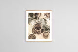 Abstract art print with beige, brown, and gray circular forms layered and textured like sand and stone, framed in light wood on a gray wall.
