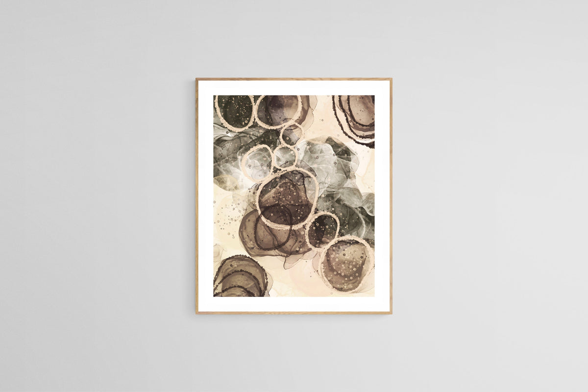 Abstract art print with beige, brown, and gray circular forms layered and textured like sand and stone, framed in light wood on a gray wall.