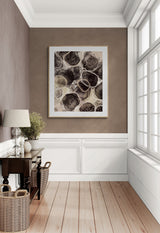 Abstract art print with circular sand-textured shapes in shades of brown and beige, framed and hung on a taupe wall in a bright, wood-floored hallway.