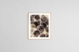 Abstract art print featuring organic circular shapes in black, brown, and beige tones with intricate textures and fluid patterns, framed in light wood and hung on a white wall.