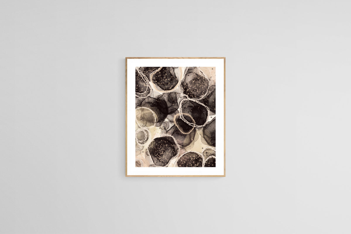 Abstract art print featuring organic circular shapes in black, brown, and beige tones with intricate textures and fluid patterns, framed in light wood and hung on a white wall.