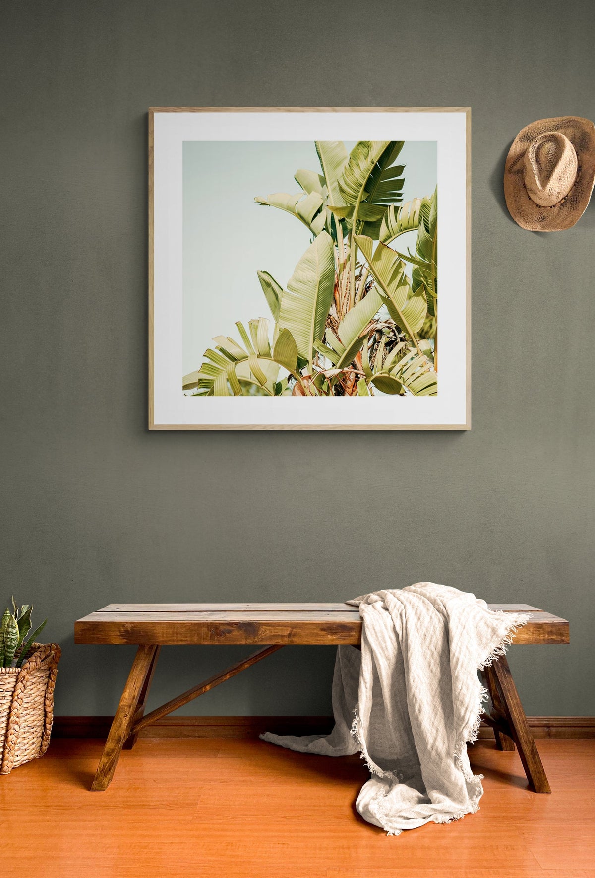 Green banana leaves against a pale sky captured in a square photo framed with light wood, hanging on a gray wall above a wooden bench with a cream throw.