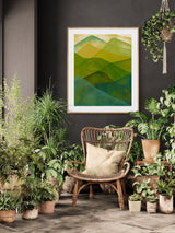 Framed abstract watercolor print of layered green mountains above a wicker chair with a beige cushion, surrounded by various potted leafy plants in a dark-walled room.