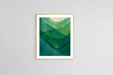 Framed abstract watercolor print of overlapping mountain shapes in gradient green shades with a muted pale circular shape above, hung on a light gray wall.