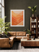 Abstract watercolor art print with layered orange and red mountain shapes framed and hung on an olive green wall above a tan leather sectional sofa.