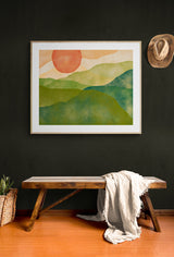 Abstract watercolor print featuring layered green hills beneath an orange sun, framed and hung on a dark wall above a wooden bench with a draped white cloth and nearby plant basket.