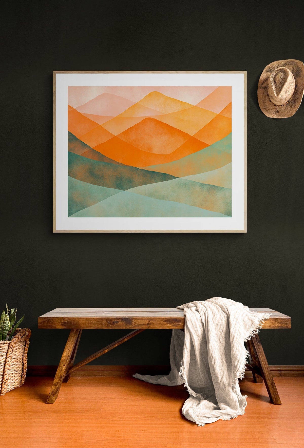 Abstract watercolor mountain landscape featuring overlapping hills in orange and green shades, framed and hung on a dark wall above a wooden bench with a draped light fabric.