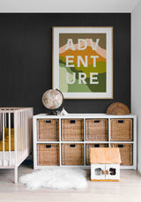 Typography art print with the word 'ADVENTURE' in large white letters over layered green, orange, and beige abstract shapes hanging on a dark wall above white storage cubes with wicker baskets