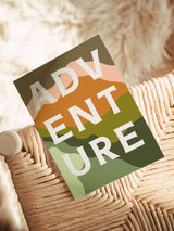 Note card with 'ADVENTURE' in large white letters over layered abstract shapes in olive green, rust orange, and peach, resting on woven wicker furniture.