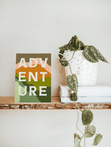 Card with stylized, layered green, orange, and pink hills and blocky white letters spelling 'ADVENTURE' on a wooden shelf beside a trailing plant in a white perforated pot.