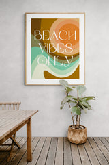 Abstract art print with layered sunset hues, teal waves, and white text reading 'BEACH VIBES ONLY' in a modern geometric font, framed on a light gray wall.
