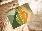 Note card with layered earth-tone abstract shapes and the phrase 'Collect Beautiful Moments' placed on woven natural fiber chair with a fuzzy cream rug in background.