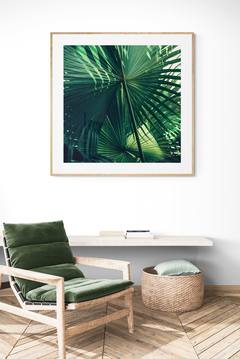 Framed photograph of overlapping green palm leaves with detailed veins, displayed above a cushioned wooden chair and woven basket in a minimalist room.
