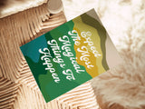 Note card with layered green, blue, and yellow abstract shapes and white cursive text reading 'Expect The Most Magical Things To Happen' placed on a woven chair seat next to a white furry rug.