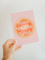 Hand holding a pink notecard with a bright orange circular gradient behind white text reading 'Focus On The Good' on a plain light background.