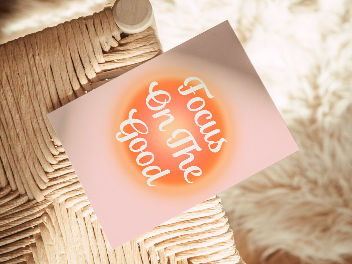 Blank notecard with a pink background and the words 'Focus On The Good' in white script centered inside an orange circular glow, placed on a woven natural fiber stool.