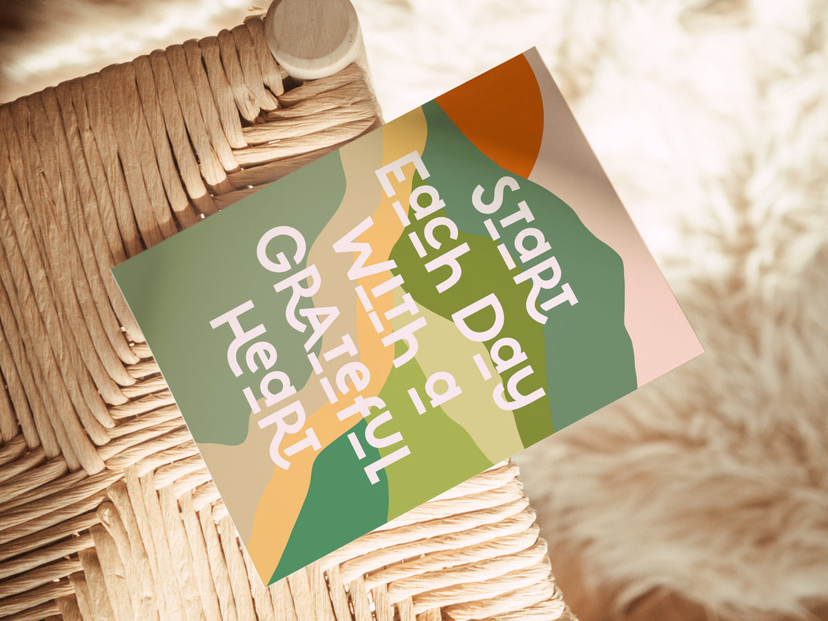 A note card with abstract green, orange, and beige shapes and the white text 'Start Each Day With a Grateful Heart,' placed on a woven, natural fiber chair seat.