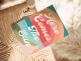 Notecard on woven seat with retro-style lettering saying 'Here Comes The Sun' in cream over layered warm rainbow arches in orange, red, green, and teal tones.
