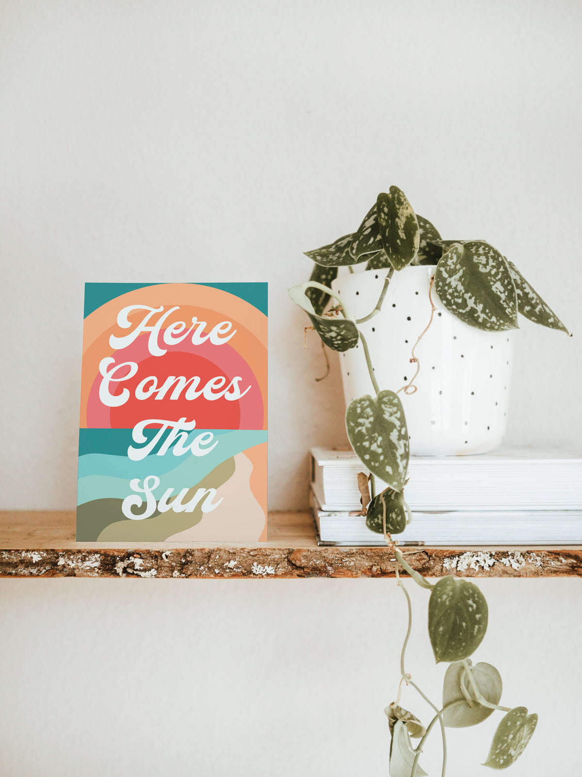 Notecard on a wooden shelf with layered waves in orange, teal, and beige and text reading 'Here Comes The Sun' in white script, next to a trailing green-leafed plant in a white pot.