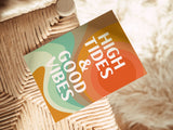 A note card with retro-inspired abstract waves in orange, green, and beige, featuring bold white text reading 'HIGH TIDES & GOOD VIBES' placed on a woven seat.