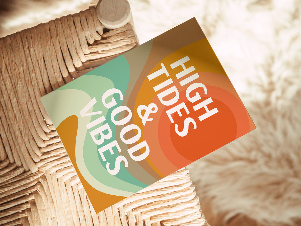 A note card with retro-inspired abstract waves in orange, green, and beige, featuring bold white text reading 'HIGH TIDES & GOOD VIBES' placed on a woven seat.
