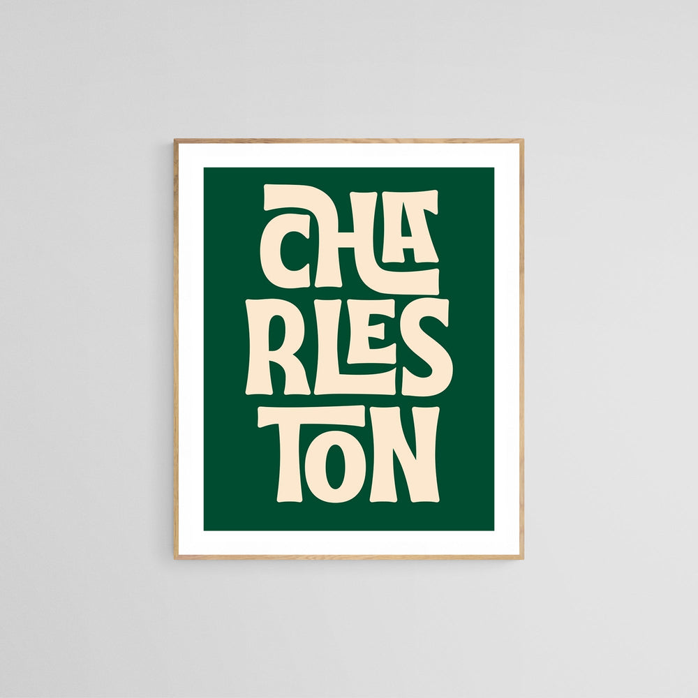 Modern art print with stylized cream text reading 'CHARLESTON' on a dark green background, framed in light wood and displayed on a light gray wall.