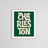 Modern art print with stylized cream text reading 'CHARLESTON' on a dark green background, framed in light wood and displayed on a light gray wall.