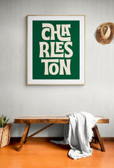 Green and cream modern art print with stylized block letters spelling 'CHARLESTON' in a geometric arrangement, hung above a rustic wooden bench with a white throw.