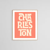 Modern art print with the word 'CHARLESTON' in bold, stylized cream letters on a coral background, framed in light wood and displayed on a gray wall.