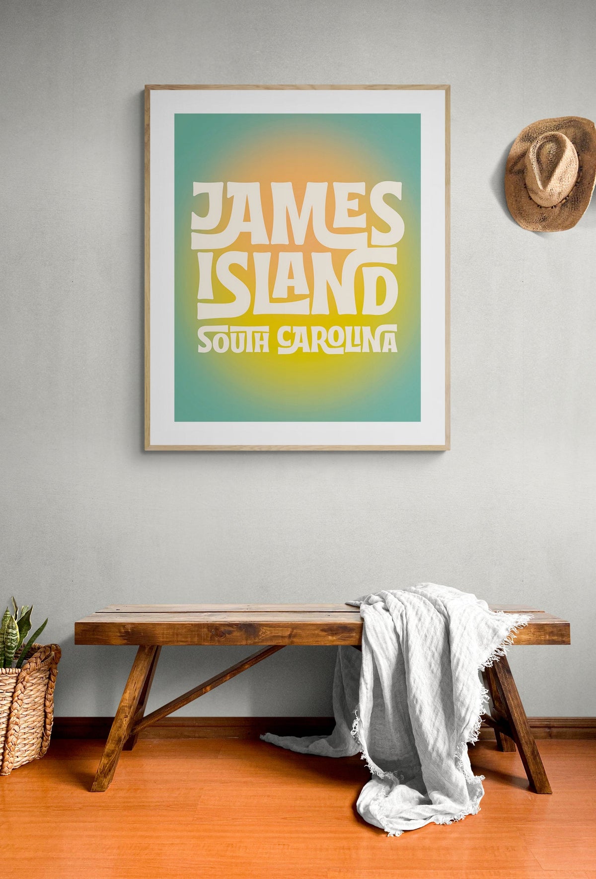 Framed modern art print with stylized white text reading 'James Island South Carolina' against a gradient background of teal and yellow, hung above a wooden bench with a white blanket and hanging straw hat.