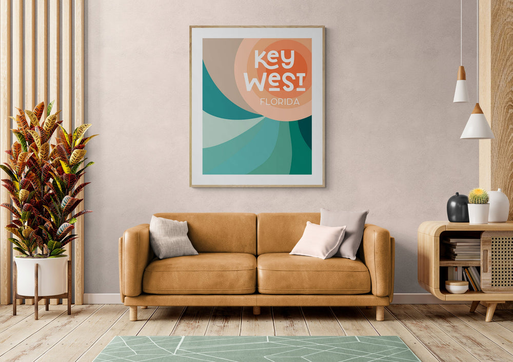 Modern living room with a tan leather sofa, decorative pillows, a large green geometric rug, potted plant on the left, wooden cabinet on the right, and colorful Key West Florida art print above.