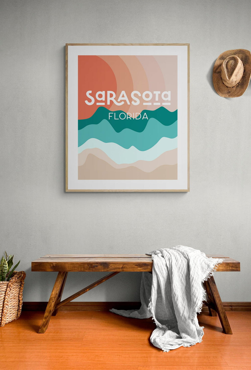 Framed modern art print with abstract wavy bands in orange, teal, and beige shades, featuring stylized text 'Sarasota Florida' on a gray wall above a wooden bench and hat.