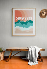 Framed modern art print with abstract wavy bands in orange, teal, and beige shades, featuring stylized text 'Sarasota Florida' on a gray wall above a wooden bench and hat.
