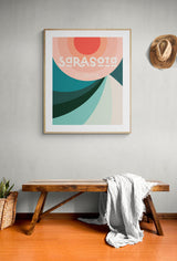 Framed modern art print of Sarasota, Florida with abstract curved shapes in shades of red, coral, teal, and turquoise above a wooden bench with a gray throw blanket, and a hat on the wall.