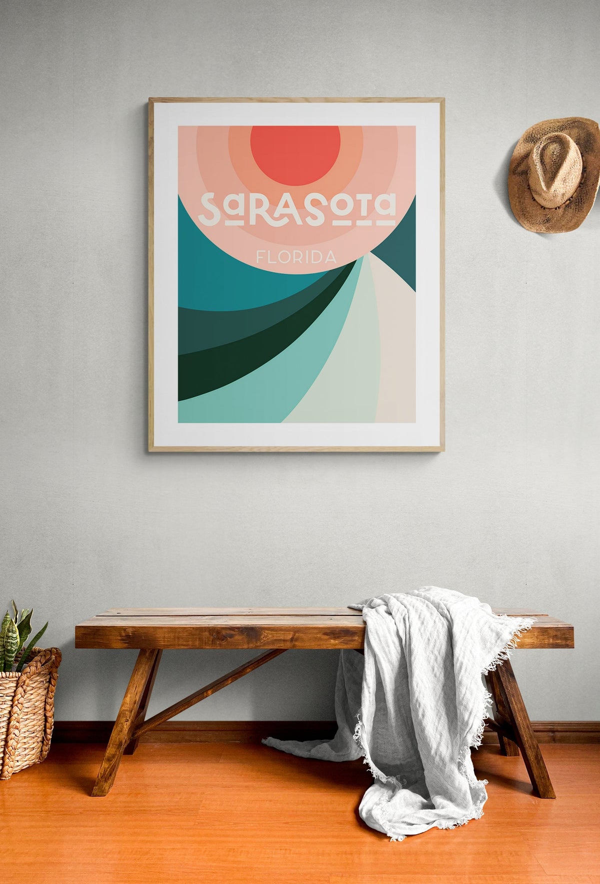 Framed modern art print of Sarasota, Florida with abstract curved shapes in shades of red, coral, teal, and turquoise above a wooden bench with a gray throw blanket, and a hat on the wall.