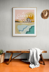Modern framed art print with stylized sandy beach, ocean waves, sun, and seagulls in muted pastel colors, labeled 'Cannon Beach, Oregon' on a light gray wall above a wooden bench.