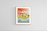 Modern art print featuring layered warm orange, green, and gray abstract shapes with bold white text reading 'Dana Point California' in a light wood frame on a gray wall.