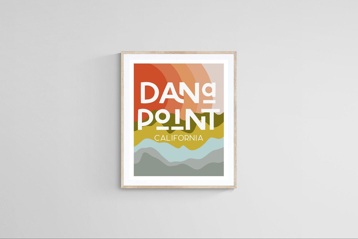 Modern art print featuring layered warm orange, green, and gray abstract shapes with bold white text reading 'Dana Point California' in a light wood frame on a gray wall.