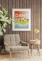 Modern Dana Point, California art print with layered red, orange, green, and blue hills, hanging on wood-paneled wall above beige fabric armchair and round wooden side table with brass lamp.