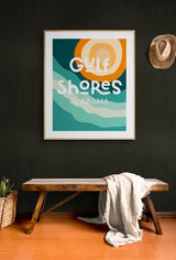 Framed modern art print with abstract turquoise waves and an orange sun, featuring stylized white text reading Gulf Shores Alabama, hung on a dark green wall above a wooden bench with a white cloth and basket nearby.