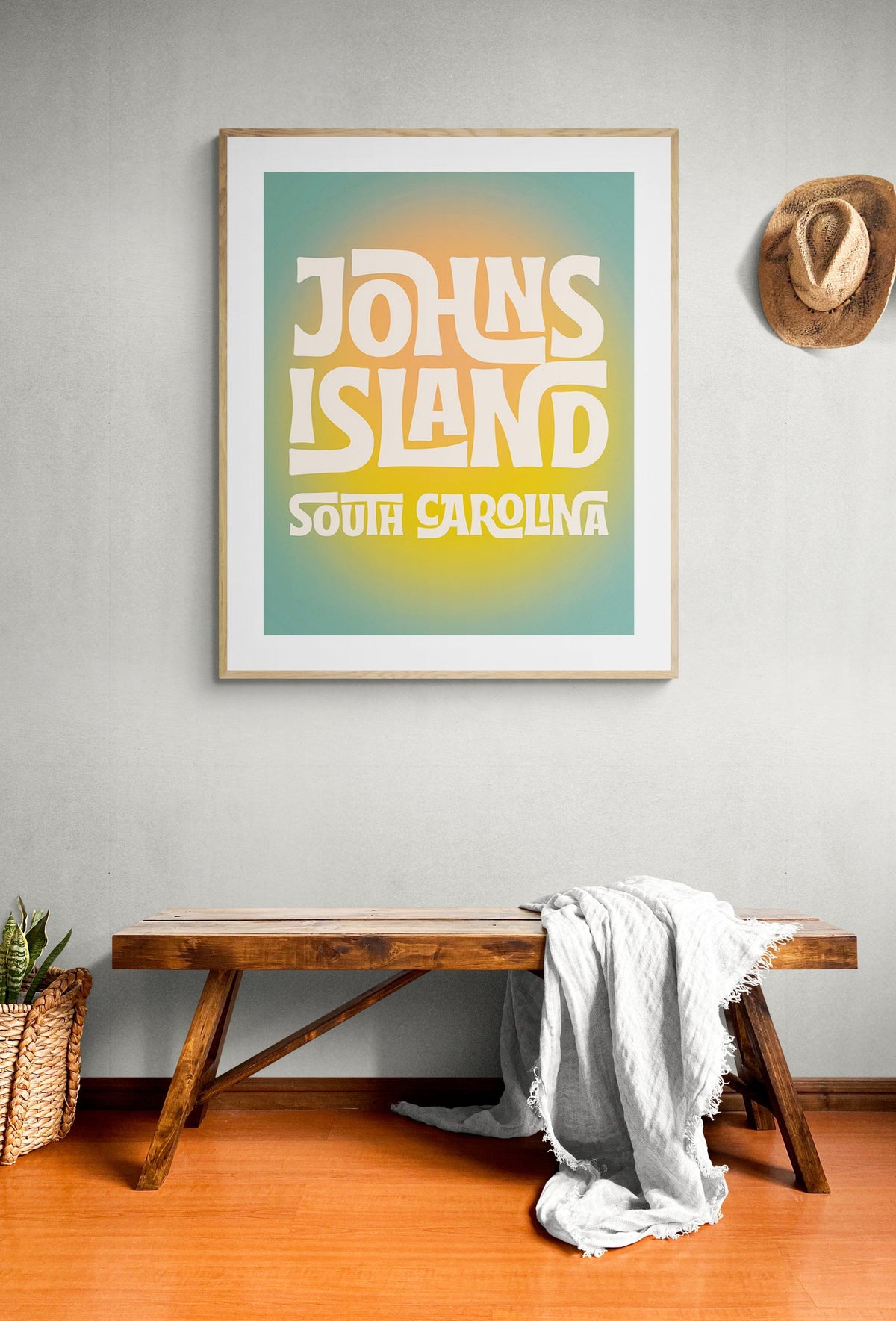Framed art print with bold, white, stylized text reading 'Johns Island South Carolina' on a gradient blue-to-yellow background, hanging above a rustic wooden bench with a white cloth draped over it.