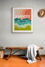 Framed modern art print featuring layered abstract hills in green, teal, and yellow with a large red sun and text 'MANTEO North Carolina' on a light wall above a wooden bench with a draped white blanket.