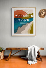 Framed modern art print depicting abstract layered landscape in earth tones with text 'Manzanita Beach Oregon' on a light wall above a wooden bench with a white throw and woven basket, hat on wall.