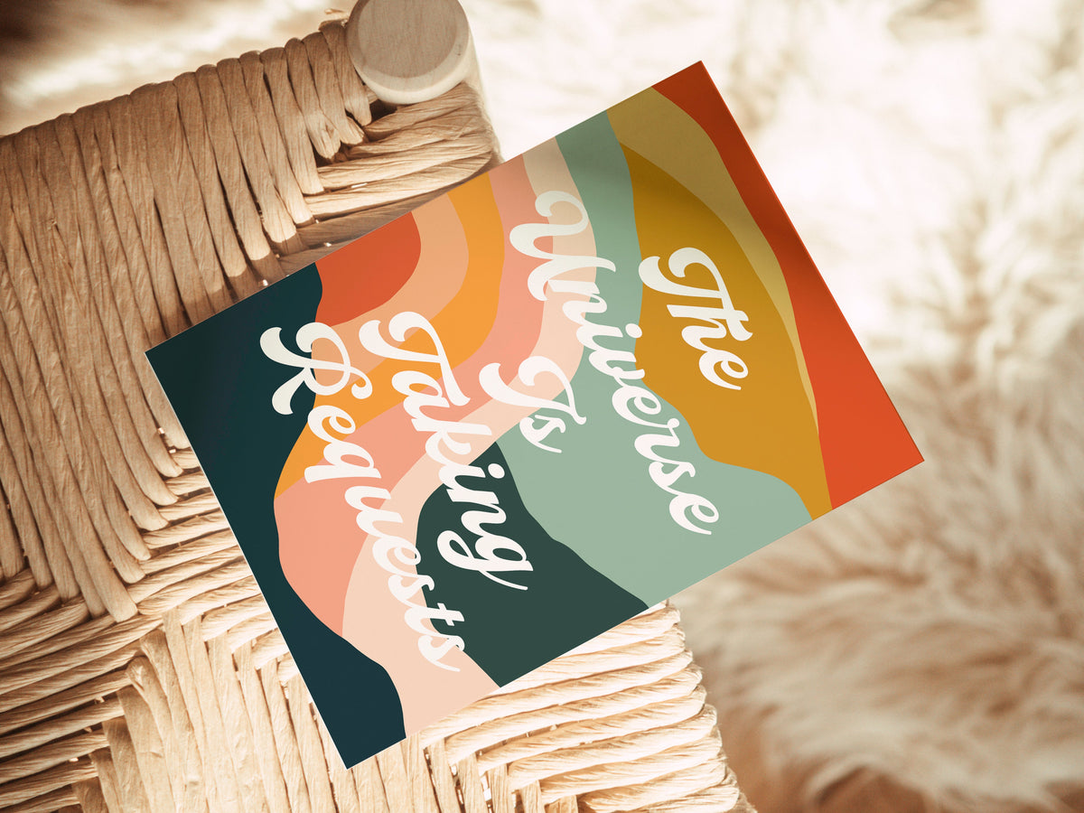 Colorful note card with layered abstract shapes in orange, pink, green, and navy, featuring cursive text "The Universe Is Taking Requests" on woven chair.