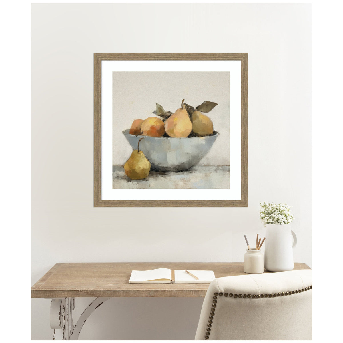 Framed watercolor painting of pears and oranges in a gray bowl with one pear outside, hanging above a wooden desk with a notebook and white vase.