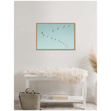 Framed canvas of a pelican row flying in a V-formation against a clear blue sky, hung on a white wall above a white bench draped with a fluffy beige throw.
