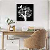 Black and white linocut art of a stylized white tree with curved branches and leaves, and a small bird perched near the top left inside a square frame, hanging above a wooden desk.