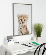 Framed portrait of a young cheetah with soft fur and dark tear markings on a white wall above a white desk with papers, a green plant, and a small owl figurine.
