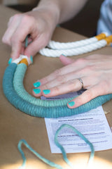 Hands with turquoise nail polish wrapping teal-colored yarn around thick rope cords, with white rope cords and instructions on a table.